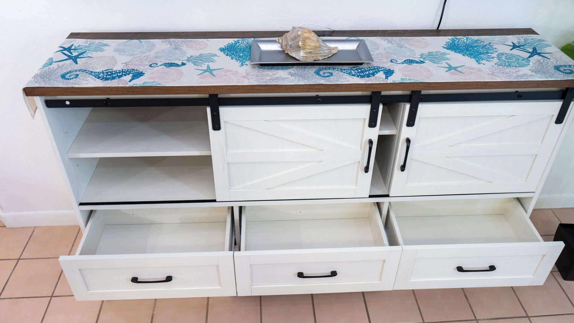 Un meuble blanc à deux portes et trois tiroirs, orné d’un dessus décoratif avec un motif en forme de coquillage et des motifs bleus.