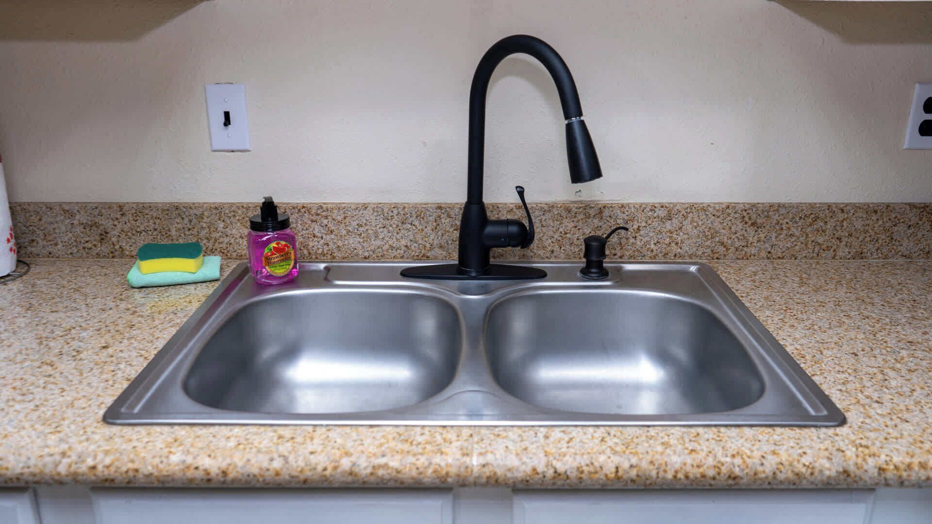 A kitchen with a double stainless steel sink, a black faucet, a bottle of strawberry jam, and a green sponge on a granite countertop.