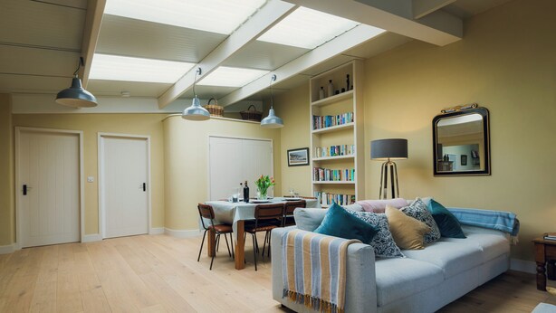 A spacious living room with a dining area, a sofa with cushions, a bookshelf, a lamp, and a mirror on the wall. - Cosy barn in Devon.Set within wonderful countryside- 15 mins from Jurassic coast (Axminster)