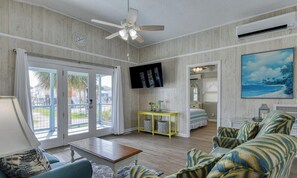 A spacious living room with a ceiling fan, a television, a yellow side table, and a large window with a view of the outdoors. - 318 Underwood Drive (South Wind 2.0)- Three Bedroom Home with a Private Pool! (Garden City Beach)