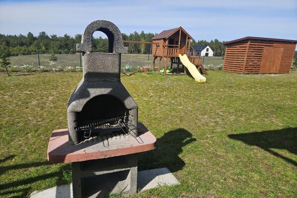 A stone-built outdoor grill with a chimney, a wooden playhouse with a slide, and a fenced yard. - Comfortable Holiday Home With a Terrace and Garden (Karsin)