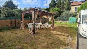 Un espace extérieur abrité, doté de tables et de chaises, d’une structure en forme de pergola et d’un véhicule blanc garé à proximité.