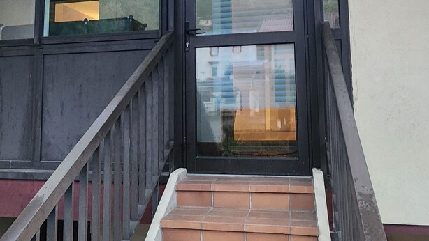 Un escalier recouvert de carreaux de terre cuite mène à une porte en verre qui offre une vue sur un immeuble et un ciel dégagé.