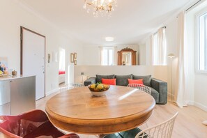 A modern living room with a round wooden dining table, a grey sofa with red cushions, a large mirror, and a chandelier.