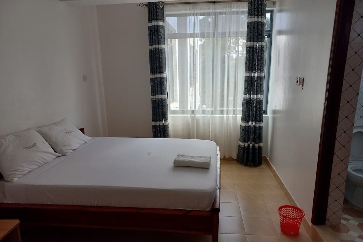Une chambre à coucher avec un lit, une tête de lit en bois, une fenêtre avec des rideaux à motifs, un panier rouge et un sol carrelé.