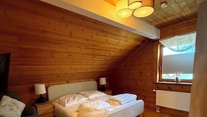 A cozy bedroom with a wooden ceiling, a bed with white bedding, a nightstand with a lamp, and a window with a view.