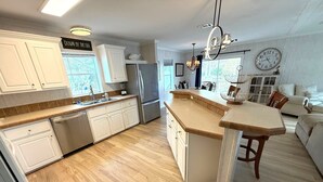 A modern kitchen with white cabinets, a built-in dishwasher, a refrigerator, a dining area with a wooden table, and a clock on the wall. - Beachside Gem Steps from Intracoastal (St. Augustine)