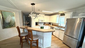 A modern kitchen with a central island, wooden chairs, a stainless steel refrigerator, and white cabinetry. - Beachside Gem Steps from Intracoastal (St. Augustine)