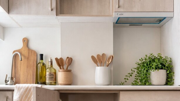 A kitchen with wooden cabinets, a countertop with a cutting board, bottles, wooden spoons, a plant, and a stove hood.