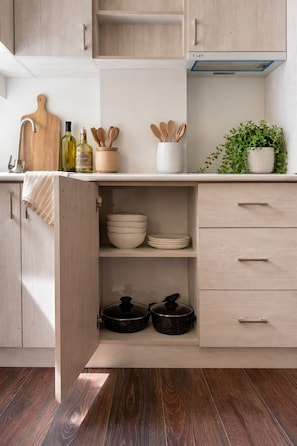 Une cuisine avec des meubles de rangement en bois, un plan de travail avec une planche à découper, des bouteilles, des cuillères en bois, une plante et une hotte de cuisinière.