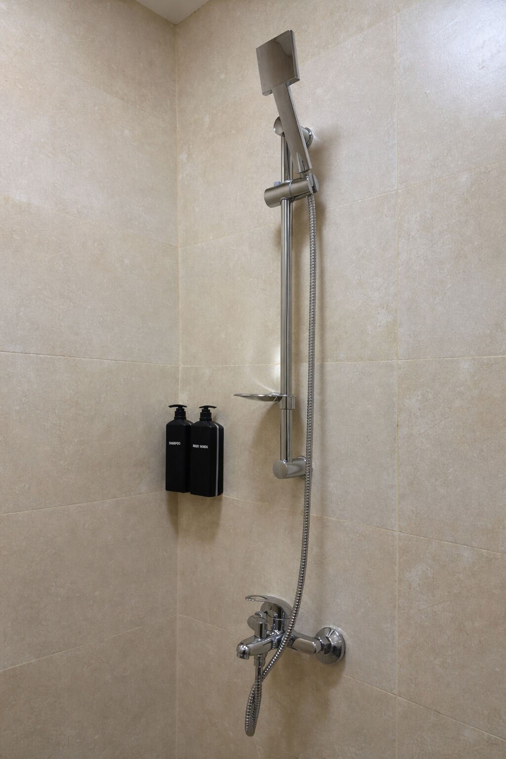 Una ducha con cabezal de ducha manual y grifo empotrado en la pared, un perchero y dos botellas de champú.