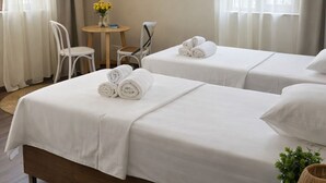A hotel room with two beds, a small table with a chair, a wall-mounted air conditioner, and a window with curtains.