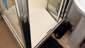 Une cabine de douche fermée par des parois en verre, avec un sol blanc, des murs recouverts de carreaux beiges et une pomme de douche visible.