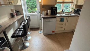 Une cuisine compacte équipée d’un lave-vaisselle, d’une machine à café et d’un évier. On y trouve une fenêtre avec des stores, une porte et un petit coin salon avec une chaise et un tabouret.