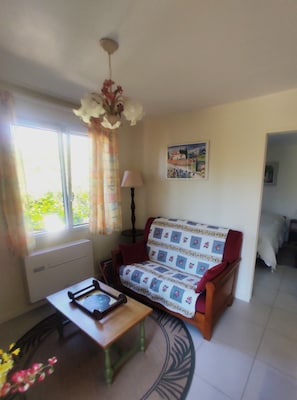 A cozy living room with a sofa, a table, and a lamp. There is a painting on the wall and a window with curtains. - Rent F1 in Luberon near 30 km from Aix en Provence, Cavaillon, (Cadenet)