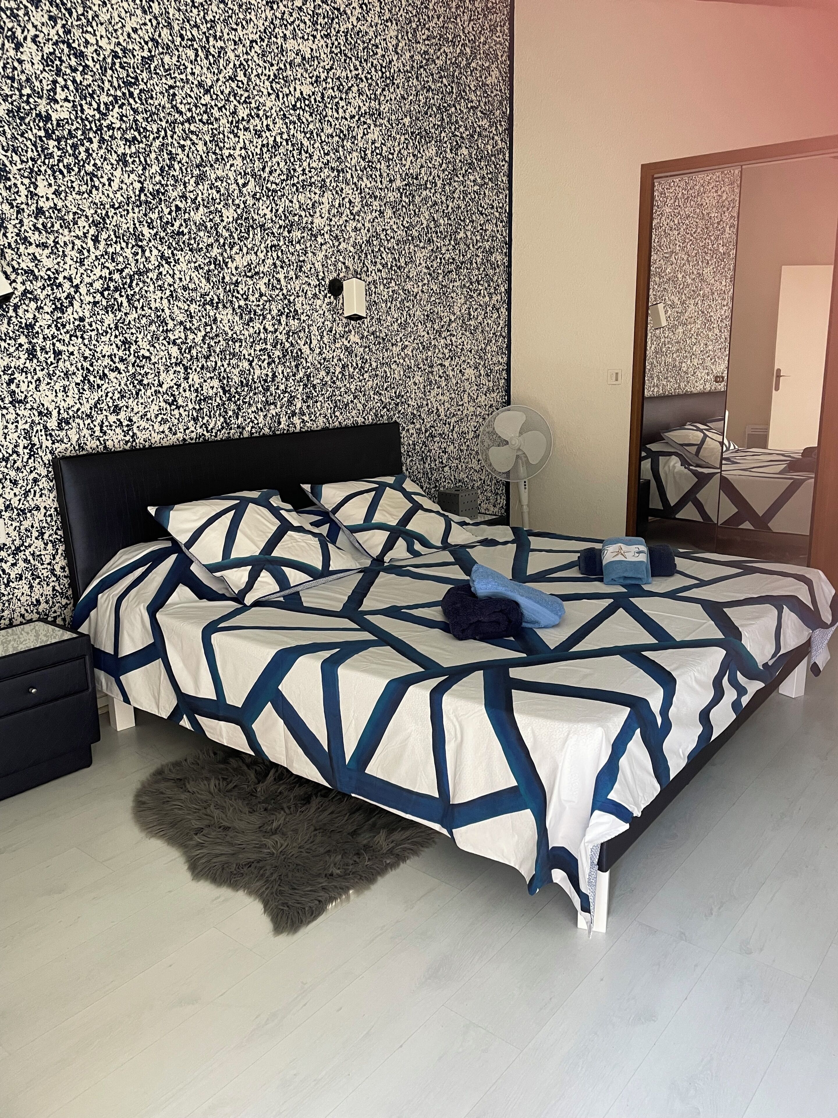 A bedroom with a bed, a nightstand, a fan, and a mirror.