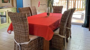 Un coin repas avec une table rouge, des chaises en osier, un vase rempli de fleurs et une vue sur l’extérieur.