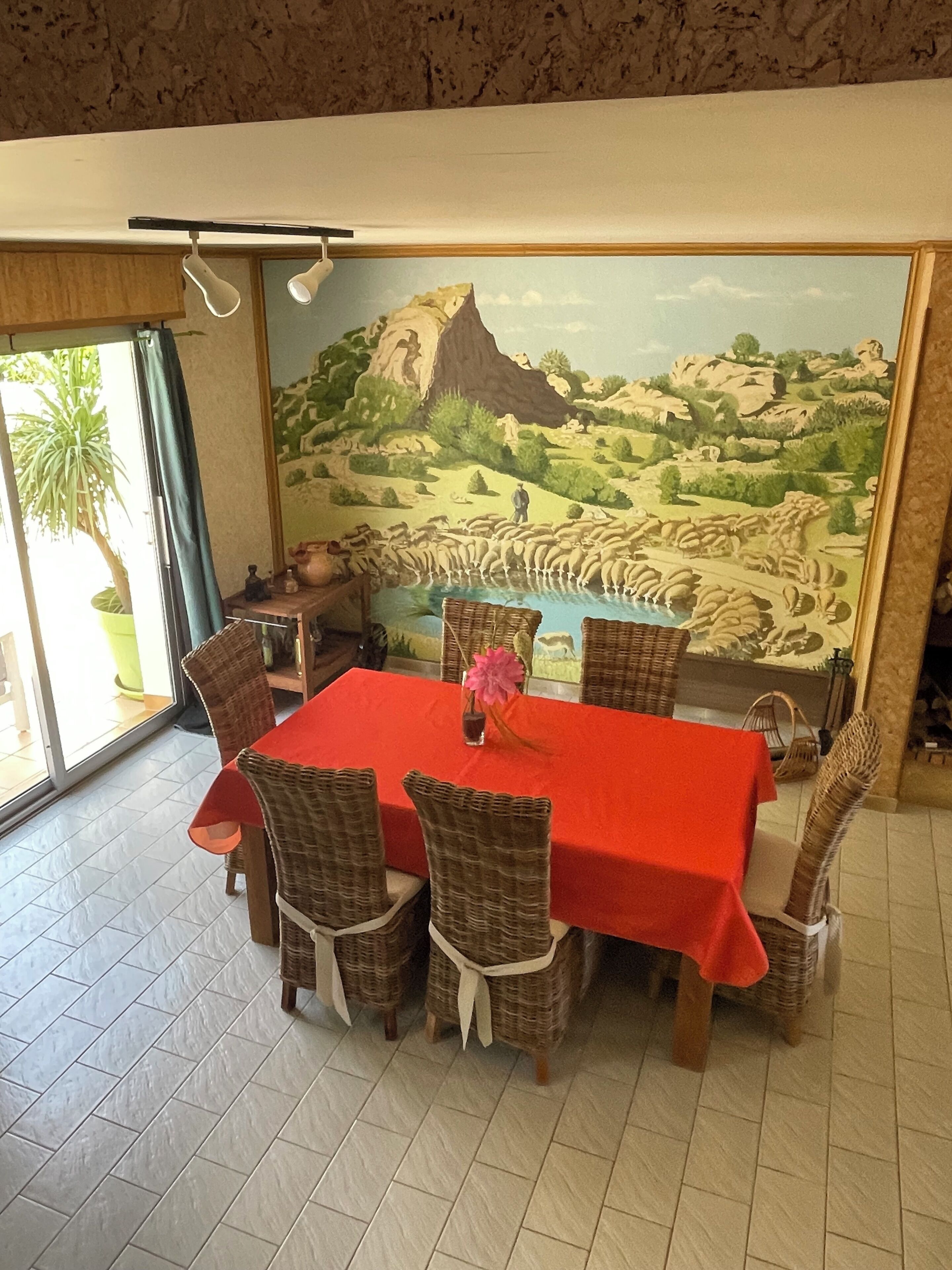 A dining area with a red table, wicker chairs, and a mural of a landscape.