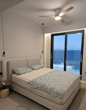 Camera da letto con un letto, una ventola a soffitto, un comodino e vista sull'oceano tramite una porta scorrevole.