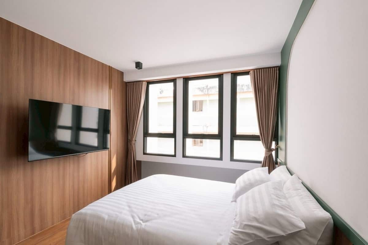Ein Hotelzimmer mit Bett, einem an der Wand befestigten Flachbildfernseher, einem Fenster mit Vorhängen und einer Fernbedienung auf einem Nachttisch.