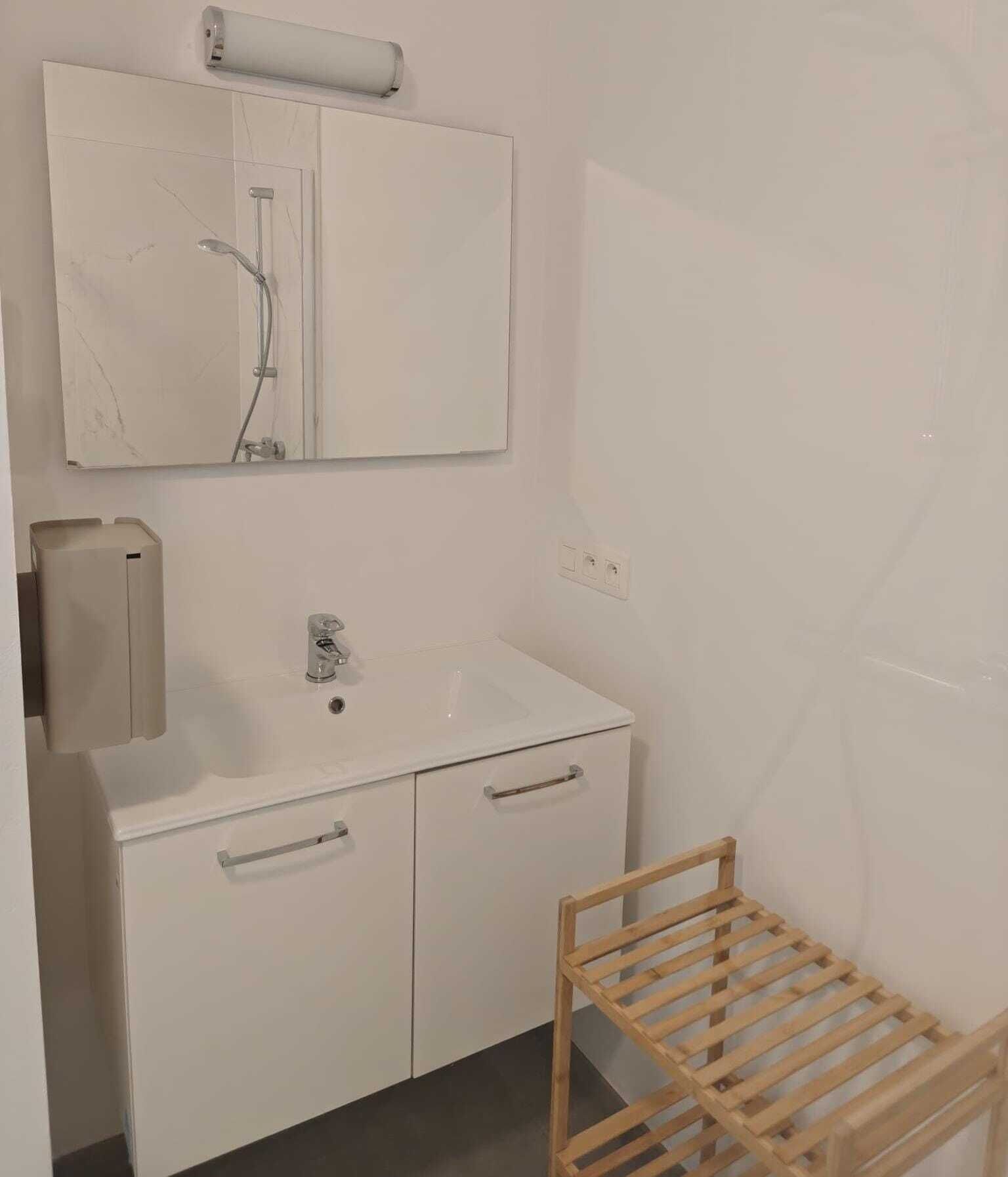 Une salle de bain avec un lavabo blanc, un miroir et une douche. On y trouve un étendoir en bois et un distributeur de serviettes.