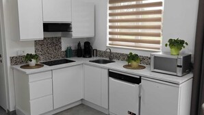 A modern kitchen with white cabinets, a dishwasher, a microwave, and a sink. There are two potted plants on the counter, and a window with blinds. - Stork country (Brugelette)