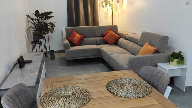 A modern living room with a grey sectional sofa, two orange cushions, a wooden dining table with woven placemats, and a small white side table with a plant. - Stork country (Brugelette)
