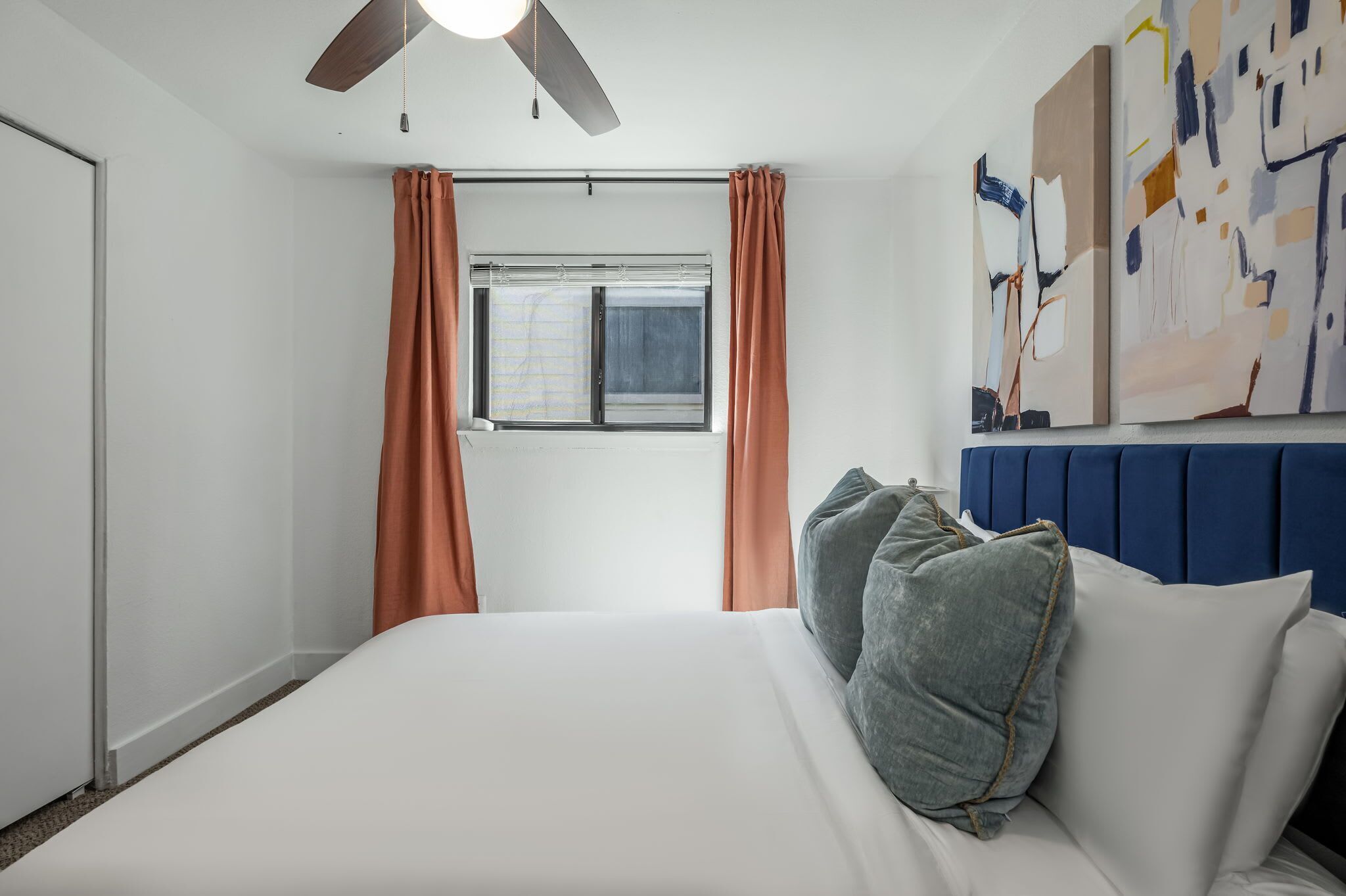 Een slaapkamer met een bed, een plafondventilator, een raam met gordijnen en een muur met abstracte kunst.