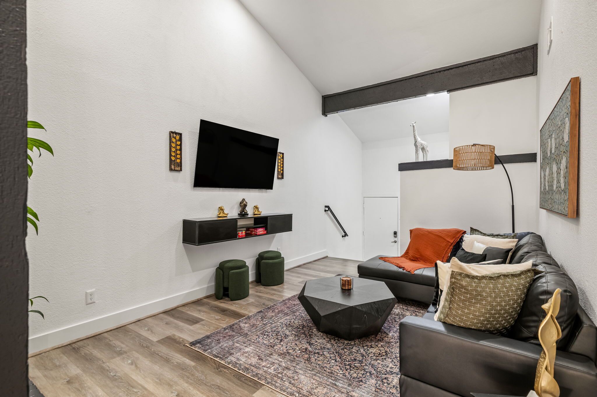 Een moderne woonkamer met een zwarte leren bank, een salontafel en een flatscreen televisie aan de muur. Er staan twee groene poefs, een kamerbreed tapijt en een hanglamp.