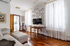 Una sala de estar con sofá, una mesa de comedor con sillas, un televisor montado en la pared y una ventana con cortinas.