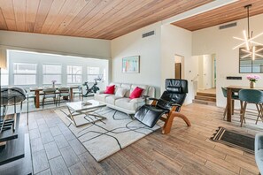 Ein geräumiges Wohnzimmer mit Holzboden, einem weißen Sofa mit roten Kissen, einem schwarzen Ledersessel und einem Essbereich mit Tisch und Stühlen.