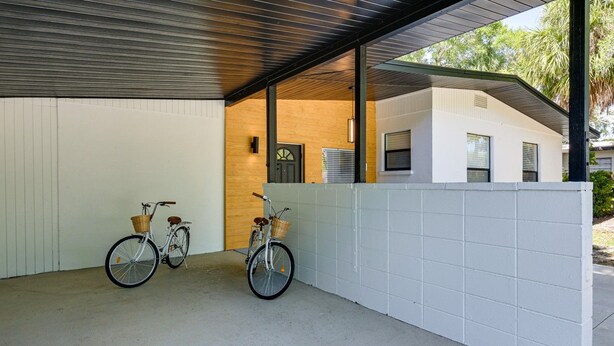 Área externa coberta com duas bicicletas estacionadas em frente a uma parede branca, uma porta de madeira e um edifício ao fundo.