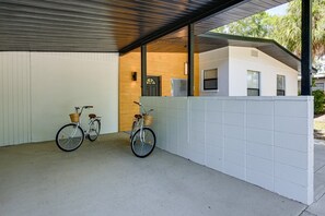 A covered outdoor area with two bicycles parked against a white wall, a wooden door, and a building in the background. - Legacy Trail Retreat  Bikes, Peloton + Fire Pit (Sarasota)