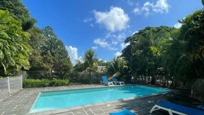 A rectangular swimming pool with clear blue water, surrounded by a stone deck. There are lounge chairs with blue cushions and a white fence enclosing the area. - Kay Ti Sable - Appartement sur la plage (Le Diamant)