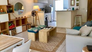 A modern living room with a dining area, a kitchen in the background, and a large window. - Kay Ti Sable - Appartement sur la plage (Le Diamant)