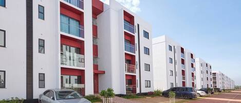 Un moderno edificio de apartamentos con varios balcones, un camino de entrada pavimentado y coches estacionados.