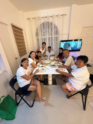 A group of people seated around a dining table with food, in a room with a TV and a window with curtains. - Casa De Amor Villa 1338 - Panglao (Dauis)