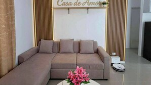 A modern living room with a sectional sofa, a round coffee table with a vase of flowers, and a wall-mounted sign that reads "Casa de Amor". - Casa De Amor Villa 1338 - Panglao (Dauis)