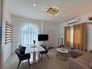 A modern living room with a dining area, featuring a large window with sheer curtains, a flat-screen TV, a sofa, and a coffee table. - Casa De Amor Villa 1338 - Panglao (Dauis)