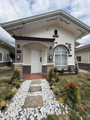 A two-story house with a white door, a stone pathway leading to the entrance, and a small garden with various plants. - Casa De Amor Villa 1338 - Panglao (Dauis)