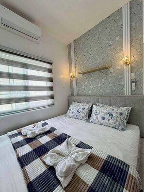A hotel room with a large bed, a window with blinds, an air conditioning unit, and a wall-mounted light fixture. - Casa De Amor Villa 1338 - Panglao (Dauis)
