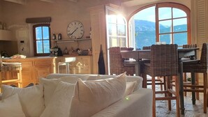 Una habitación acogedora con un ventanal grande, un comedor con sillas de mimbre, un sofá y una cocina con armarios de madera y un reloj.