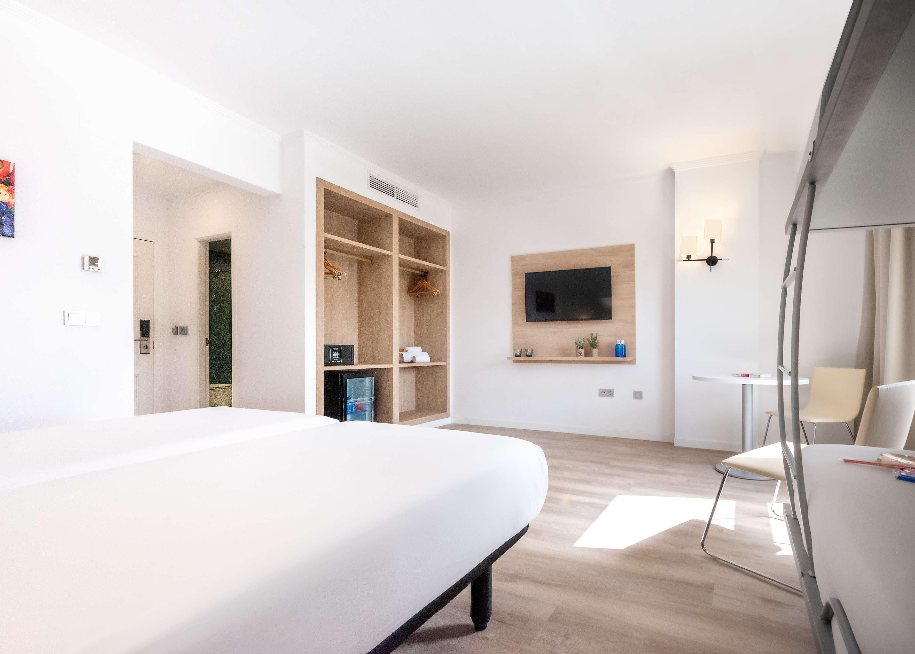 Una habitación de hotel moderna con una cama grande, armario empotrado, un televisor montado en la pared y una pequeña mesa con sillas.