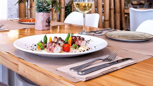 Ein Esstisch mit einem Teller Essen, einem Glas Weißwein und einer Bambuspflanze im Hintergrund.