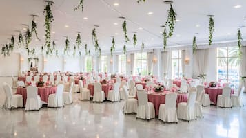 A spacious banquet hall with tables set for dining, surrounded by white chairs, and adorned with hanging greenery and string lights.