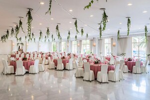 Un amplio salón de banquetes con mesas dispuestas para comer, rodeado de sillas blancas y decorado con follaje colgante y luces de adorno.
