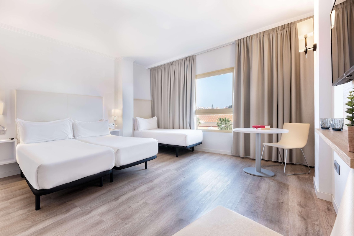 Camera d'albergo con due letti, un tavolino, una sedia e vista sulla città.