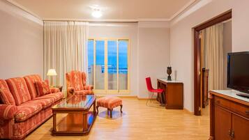 Una sala de estar con sofá, sillón, reposapiés, mesa de centro de vidrio, televisor y vistas amplias a través de un ventanal.