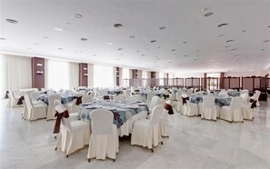 Un amplio salón de banquetes con varias mesas redondas, cada una dispuesta con sillas blancas y manteles.