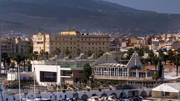 Un puerto deportivo con barcos, un edificio moderno con fachada de vidrio y un paisaje urbano montañoso al fondo.